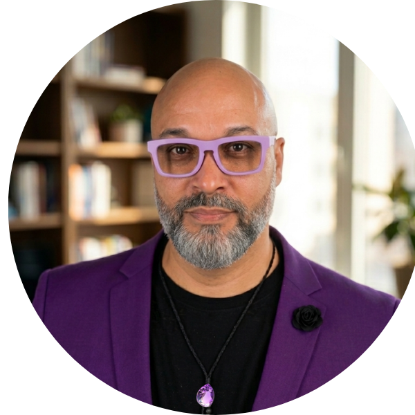 Portrait of a bald man with a gray beard wearing purple glasses and blazer, standing indoors.