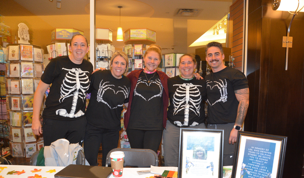 Five people in skeleton shirts posing indoors behind a table with framed signs and food.
