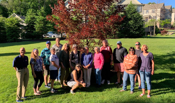 Group of people standing on a grassy lawn in front of a red-leaf tree and apartment buildings.