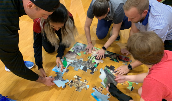 People kneeling around a floor puzzle, reaching in to fit pieces together on a wooden floor