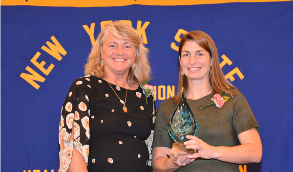 Two women holding an award trophy in front of a New York State banner