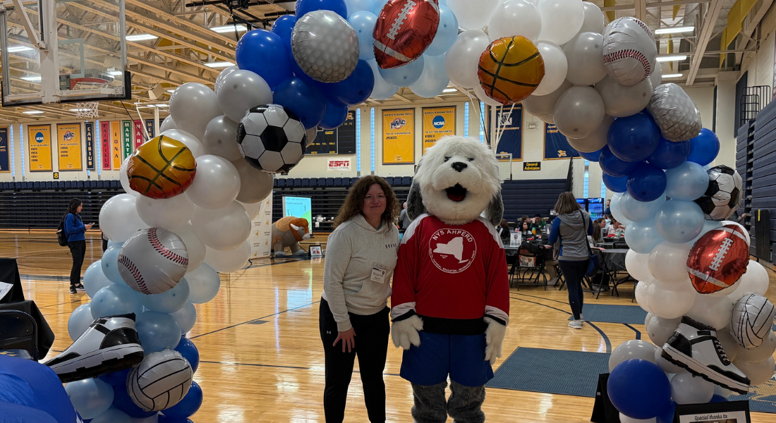 Two people standing under a balloon arch with sports balls in a gymnasium, one in a soccer mascot costume