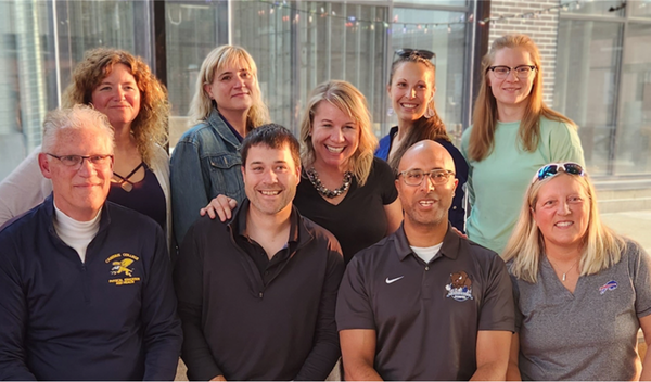 Seven smiling coworkers pose indoors in front of glass windows, wearing casual shirts and sweaters.