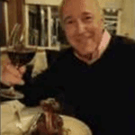 Smiling man seated at a restaurant table, holding a drink beside a dessert and bookshelves.