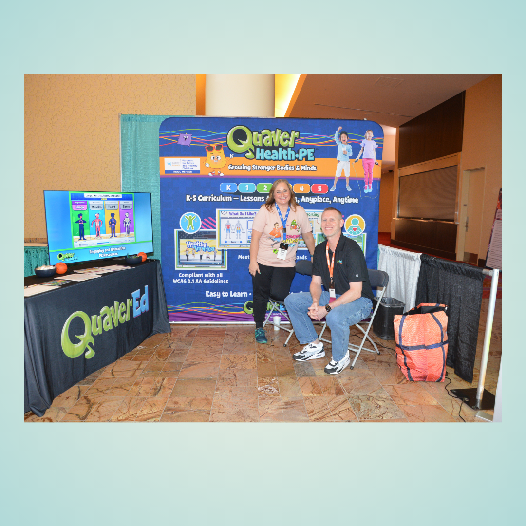 Event booth with two people in front of a Quaker Health PE display, one seated and one standing, with a TV monitor nearby