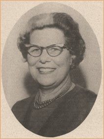 Portrait of a smiling woman wearing glasses and a necklace against a plain oval background