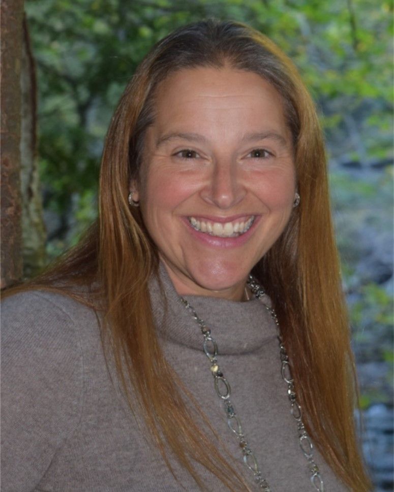 Smiling woman with long brown hair wearing a gray sweater, standing outdoors against a leafy background
