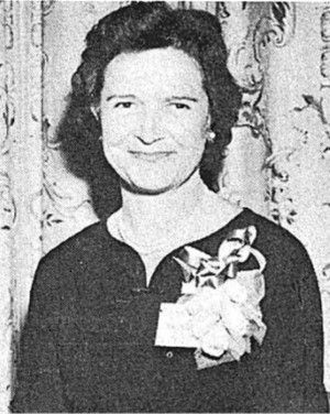 Smiling woman in a dark dress with a floral corsage, standing before patterned curtains.