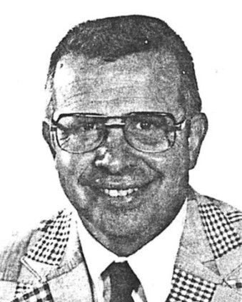 Black-and-white portrait of a smiling man wearing glasses, a checkered jacket, shirt, and tie.