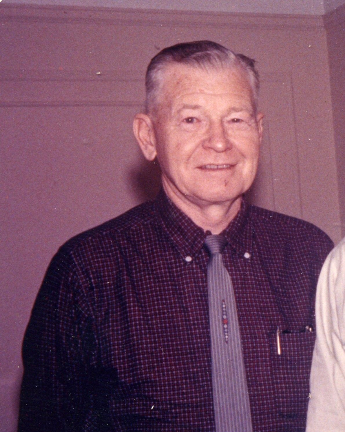 Smiling man in a dark patterned shirt and tie standing indoors against a beige wall