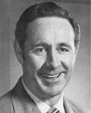 Black-and-white portrait of a smiling man in a suit and tie, facing the camera.