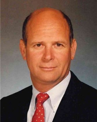 Studio portrait of a bald man in a dark suit, white shirt, and red tie against a gray background