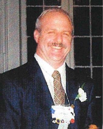 Smiling man in a dark suit and tie, wearing a white boutonniere at a formal event