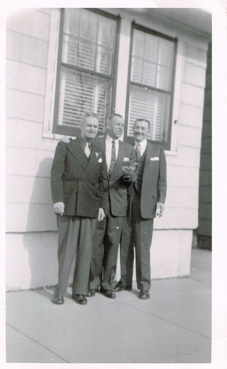 Three men in suits standing outside a building by a window and wall.