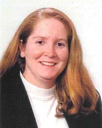 Smiling person with long red hair wearing a black blazer and white turtleneck against a white background