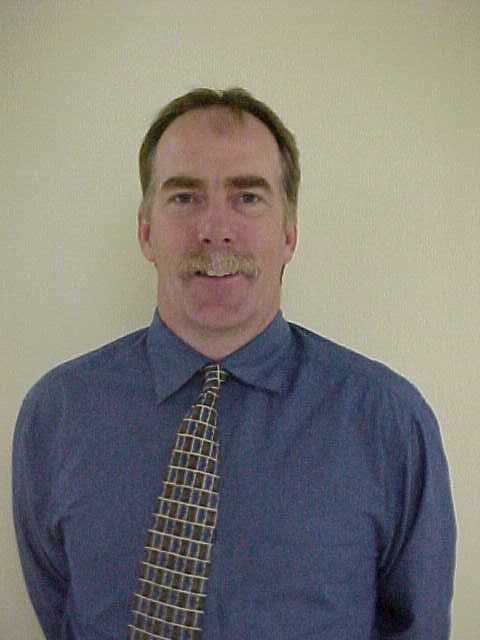 Man in blue shirt and patterned tie, standing against a plain beige wall, facing the camera.