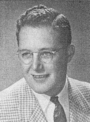 Portrait of a man in glasses and a checkered suit, smiling slightly against a plain background