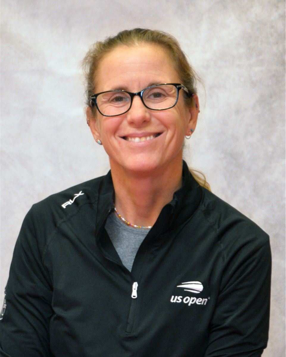 Smiling person wearing glasses and a black US Open jacket against a gray backdrop