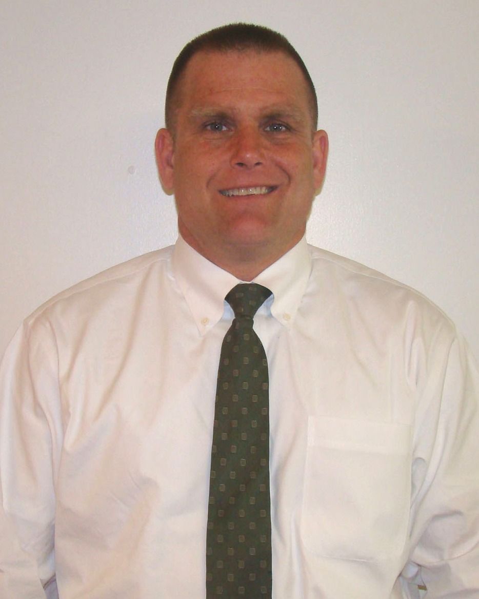 Man in a white dress shirt and dark tie, smiling against a plain light background