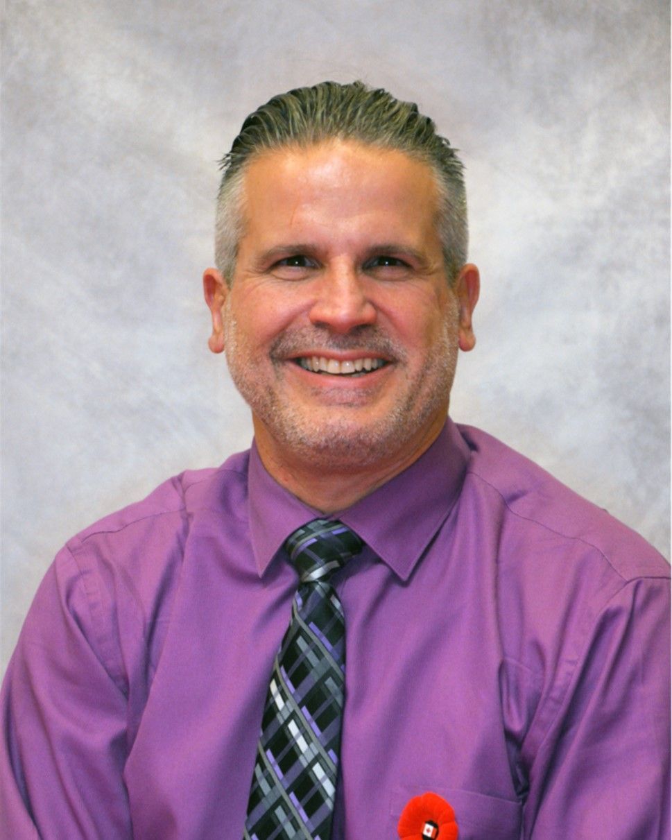 Smiling man in a purple shirt and patterned tie with a red poppy pin against a gray backdrop