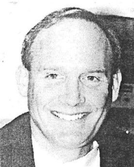 Smiling man in a dark jacket, shown in a close-up black-and-white portrait.