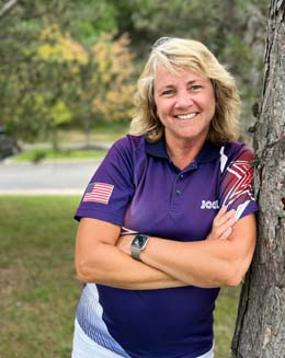Smiling person in a purple polo shirt leaning against a tree outdoors