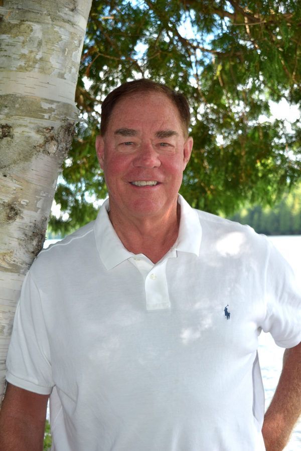 Man in a white polo shirt smiling beside a tree outdoors with greenery in the background