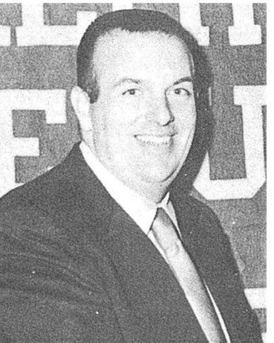 Smiling man in a dark suit and tie standing before a large patterned sign