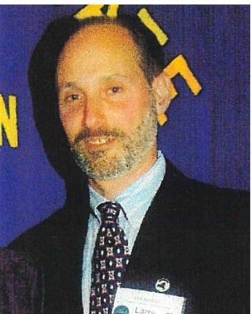 Man in a suit and patterned tie stands before a dark blue backdrop, wearing a name tag and smiling.