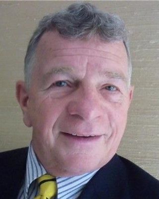 Smiling man in a suit and striped shirt, wearing a yellow tie, against a plain beige background