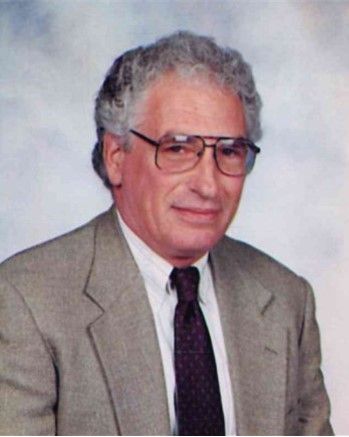 Portrait of a man in a tan suit, white shirt, and dark tie, wearing glasses, against a plain background