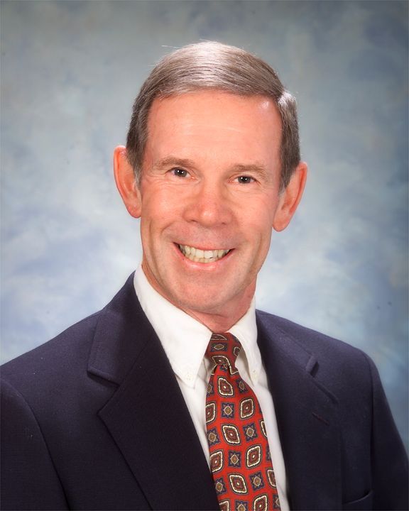 Studio portrait of a smiling man in a dark suit, white shirt, and patterned red tie against a gray backdrop