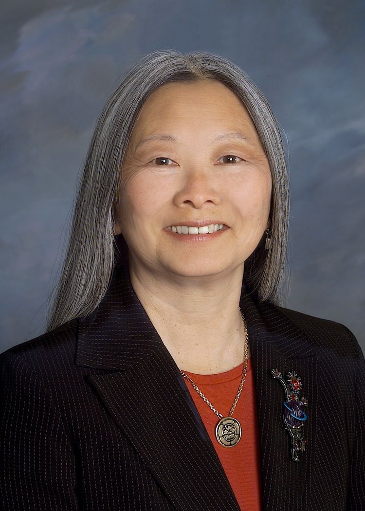 Studio portrait of a smiling woman with long gray hair in a black blazer and red top, wearing a pendant necklace