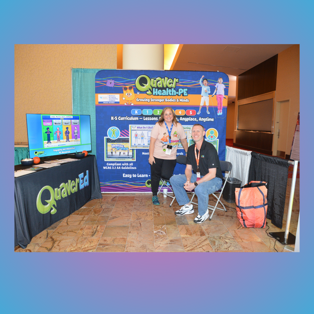 Event booth with two people in front of a Quaker Health PE display, one seated and one standing, with a TV monitor nearby
