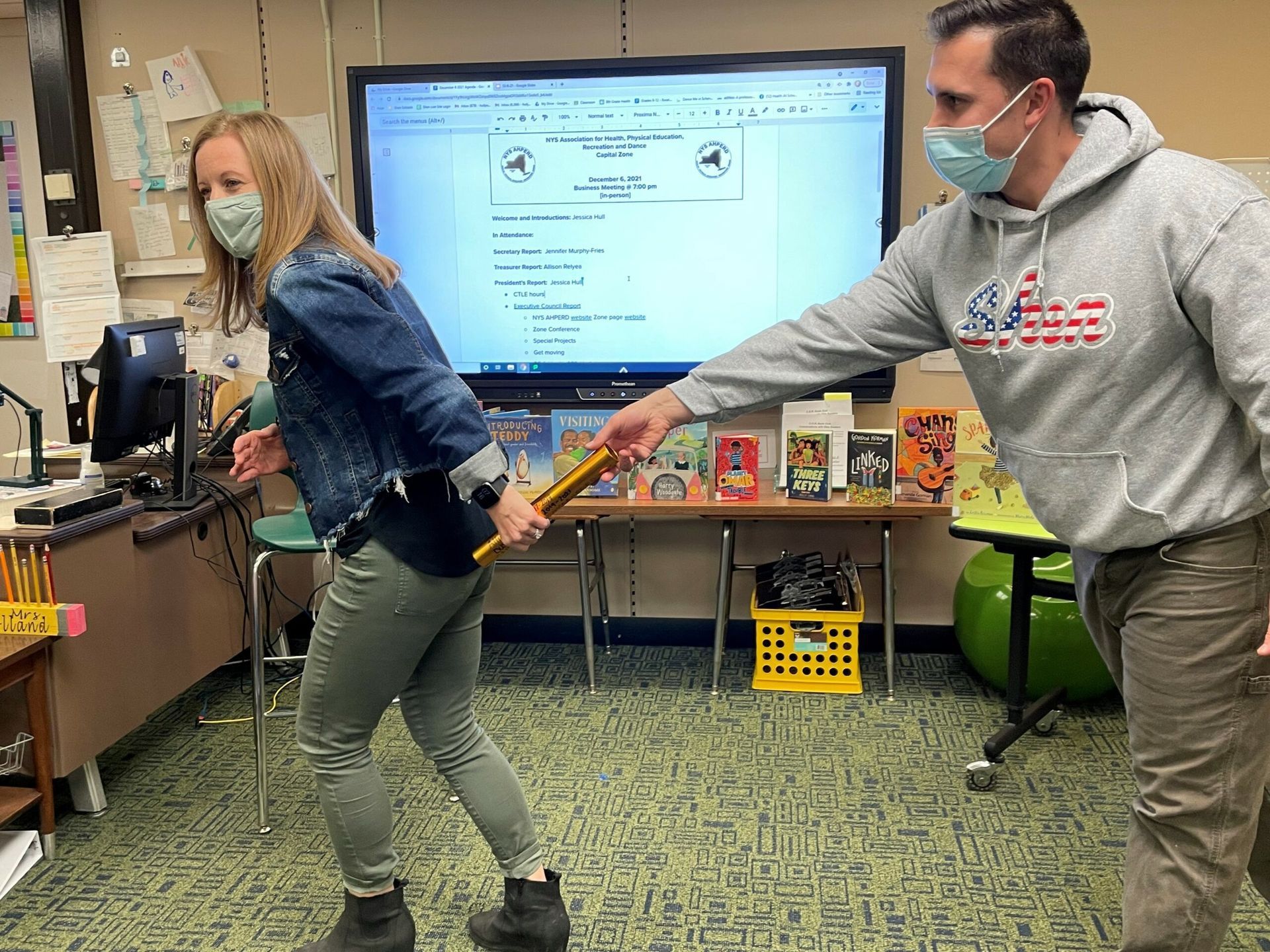 Two masked people in a classroom tug a yellow band during a hands-on activity.