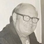 Smiling man in glasses and a suit jacket, photographed in black and white.