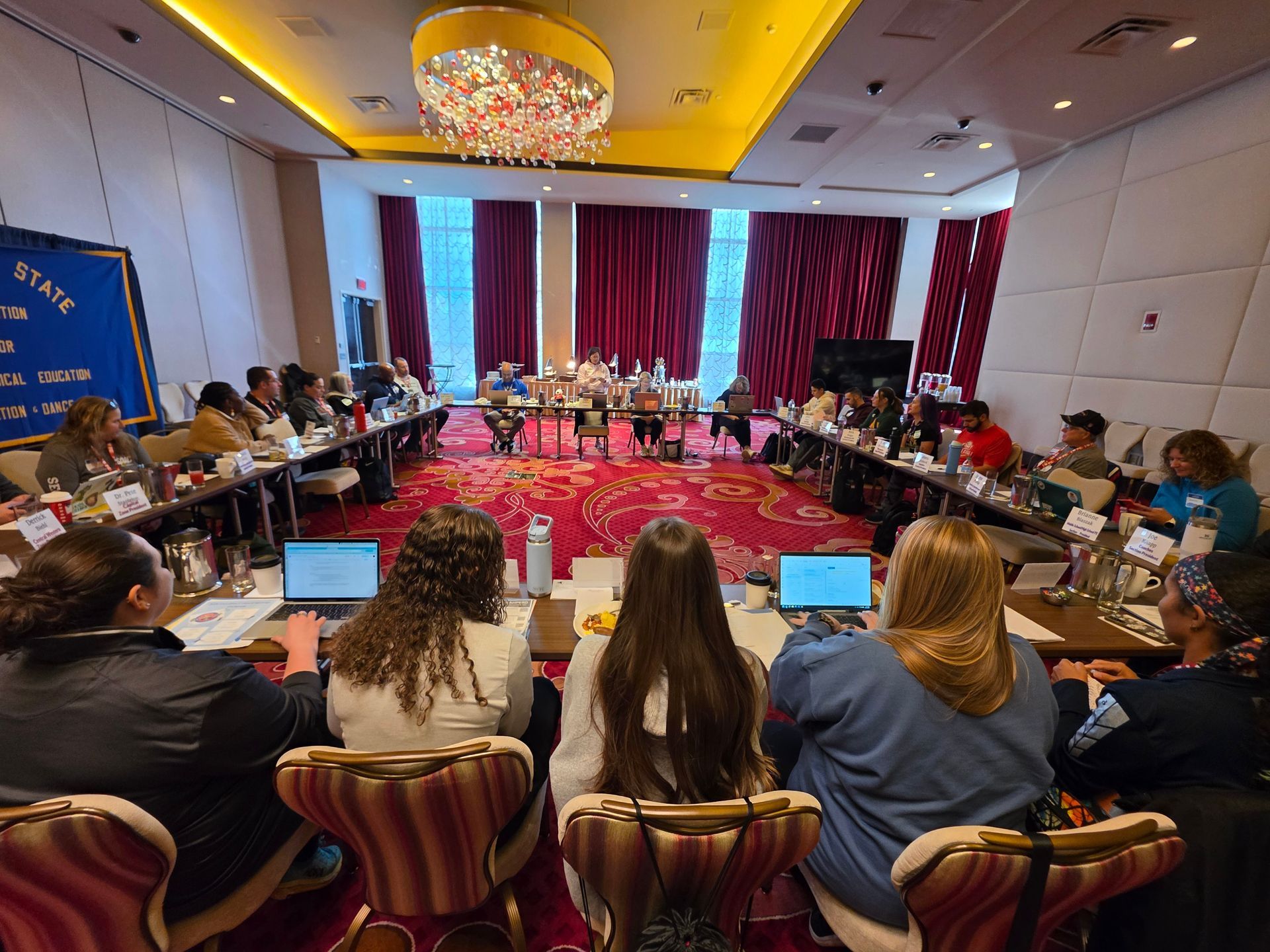 Conference room with a long roundtable, attendees seated around it, and a large chandelier overhead.