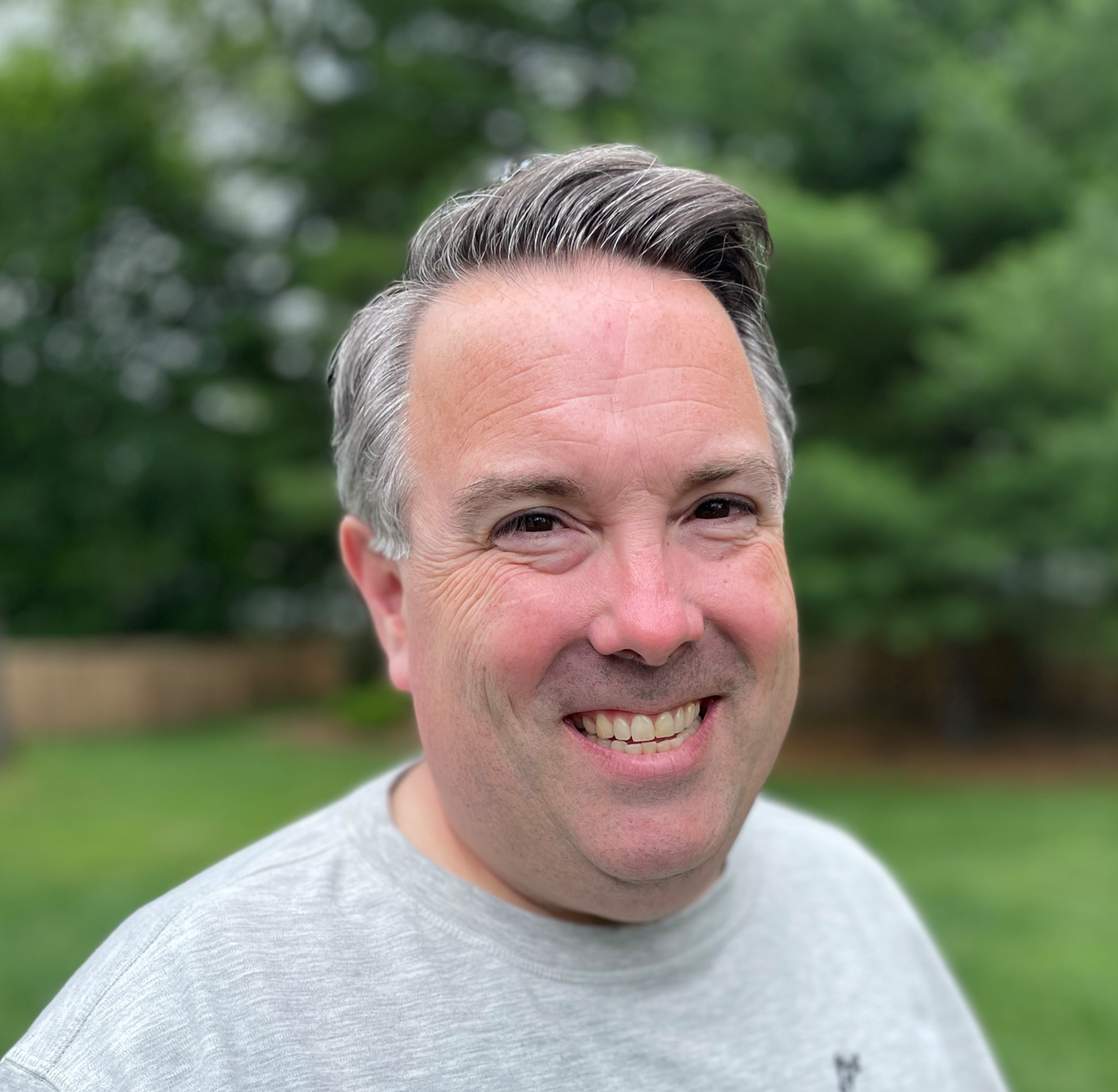 Smiling man in a gray shirt outdoors with blurred green trees in the background