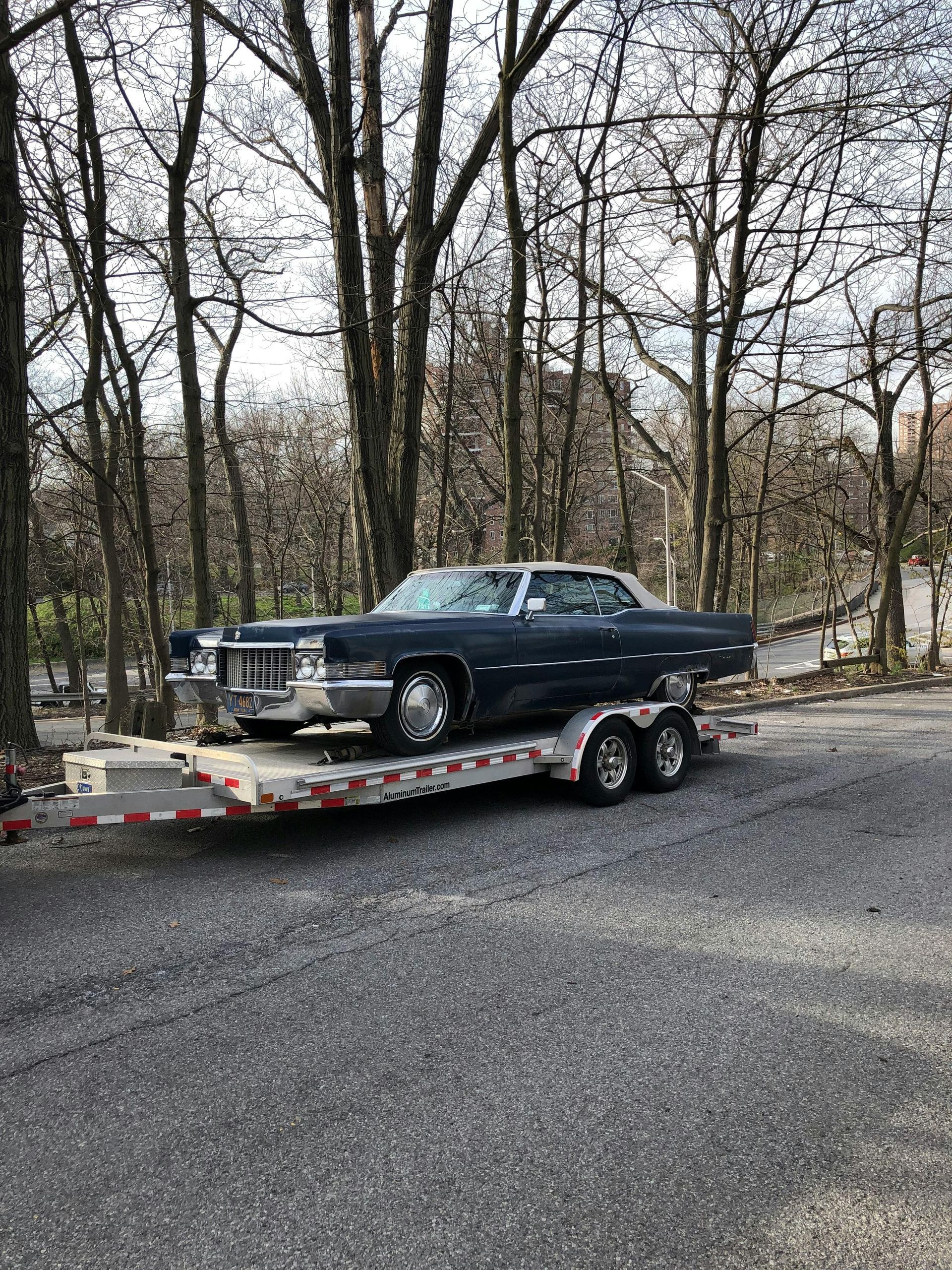Blue vintage Cadillac on a trailer, parked on a gravel road in front of trees.