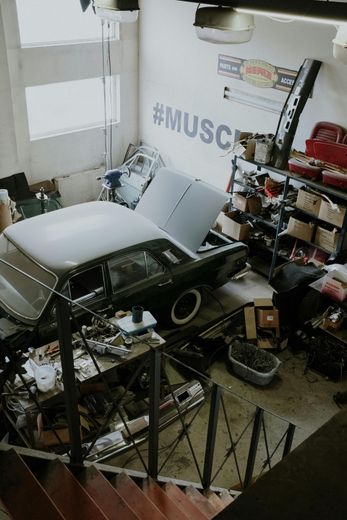 Cluttered garage with a classic car, its hood open. Tools and boxes fill the space.