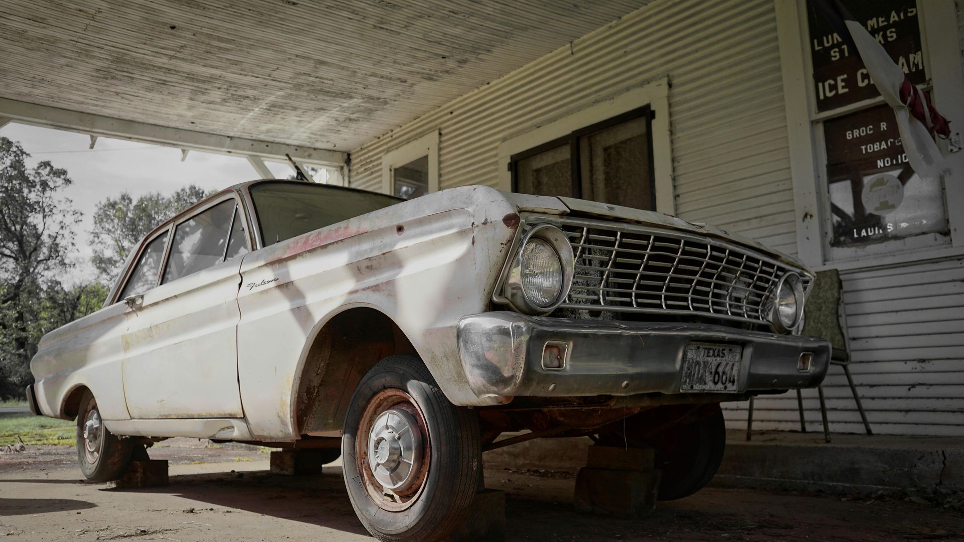 A weathered white Ford Falcon sedan parked under a porch of a rundown building.