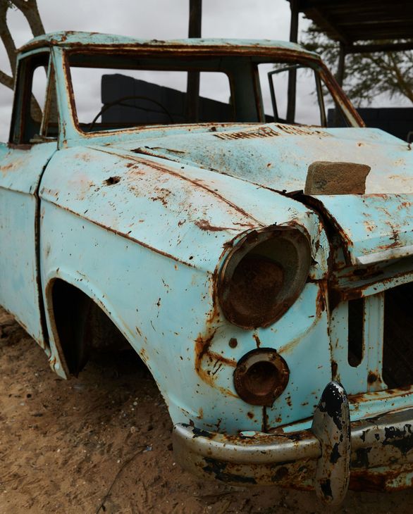 Rusted, light blue vintage pickup truck; weathered with missing headlights and exposed interior, set outdoors.