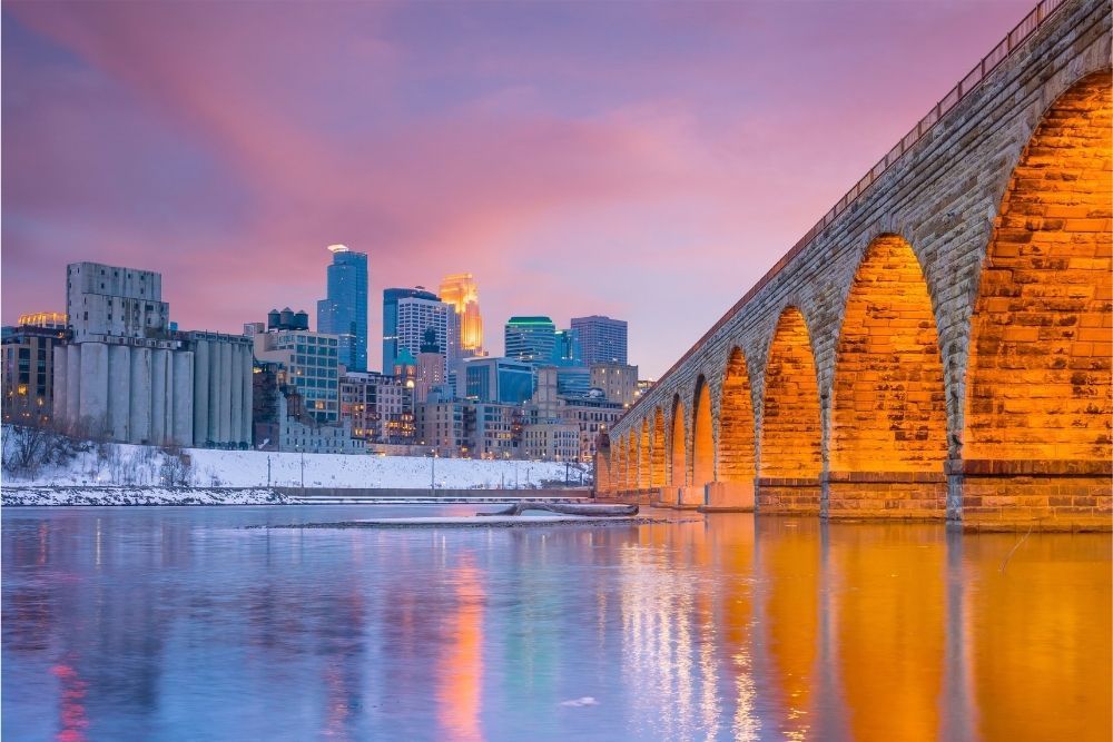 Snowy city skyline at sunset beside an arched stone bridge over calm water