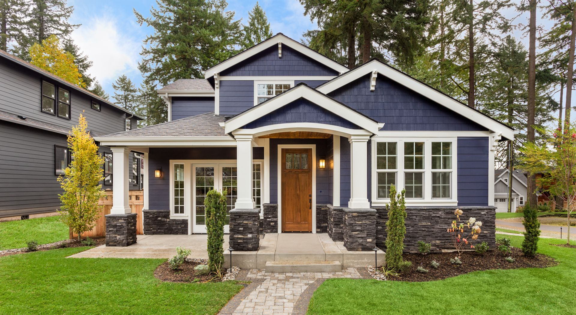 Blue craftsman-style house with white trim, stone columns, and a front porch in a wooded neighborhood