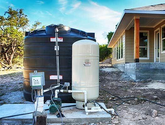 Water tank and white pressure tank beside a house, with plumbing and pump equipment outdoors.