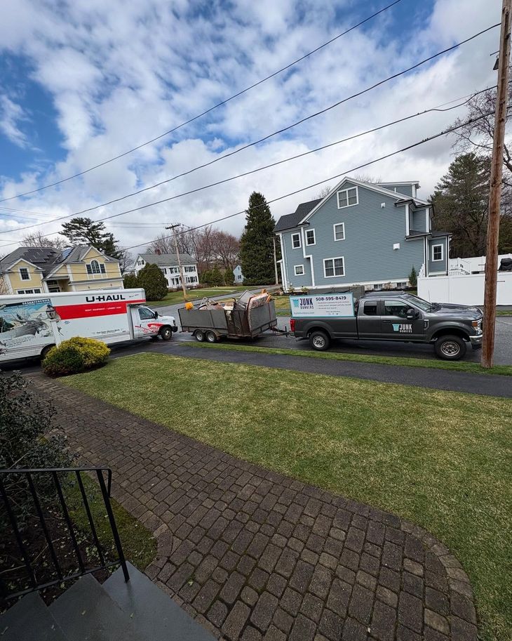 A U-Haul truck and work truck are parked on a street, with a trailer in between. Blue house in the background.