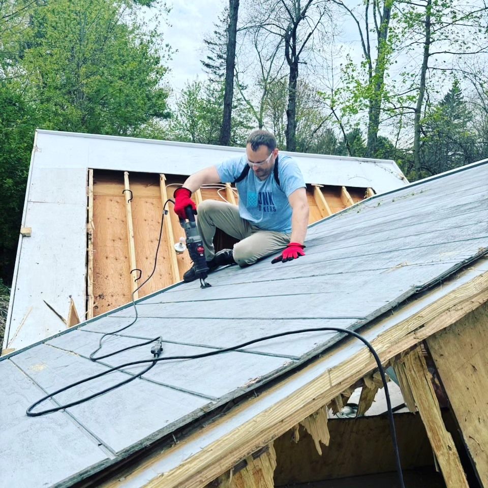 A man is kneeling on top of a roof using a drill.