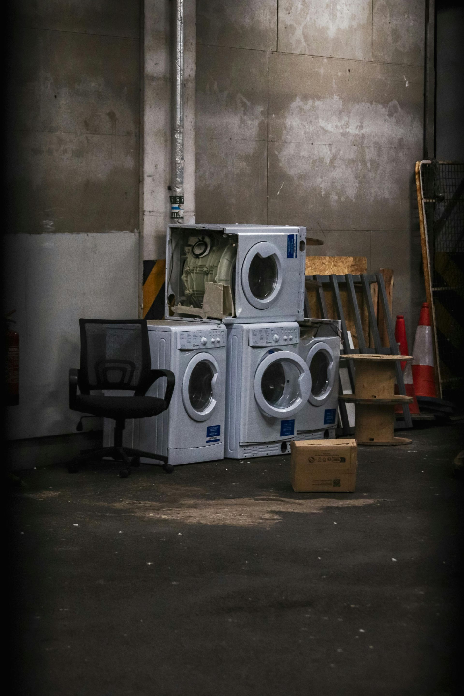 Four washing machines stacked in an industrial setting with a chair and boxes; concrete walls.