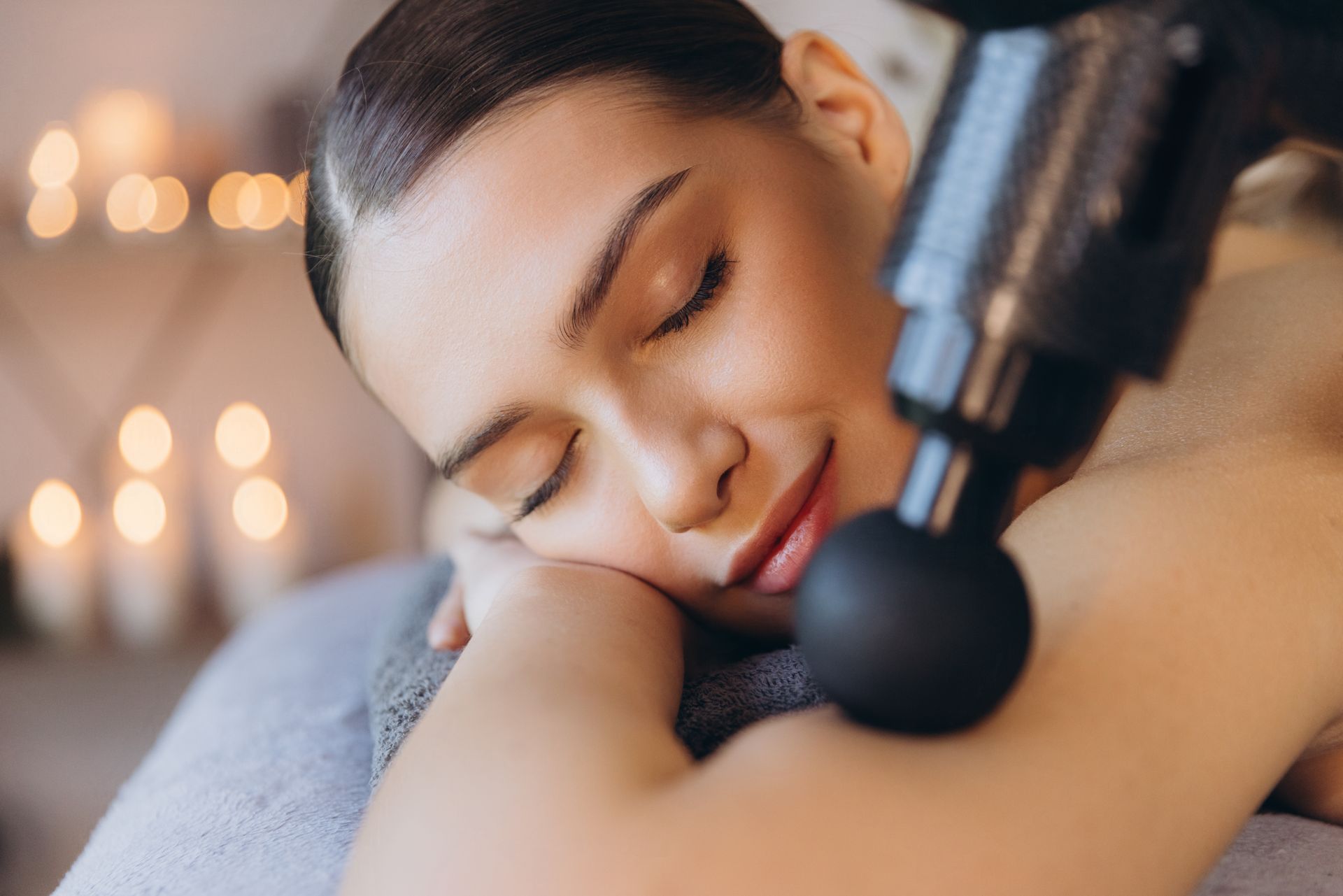 Person resting face down while a massage gun is used on their shoulder, with warm blurred lights in the background