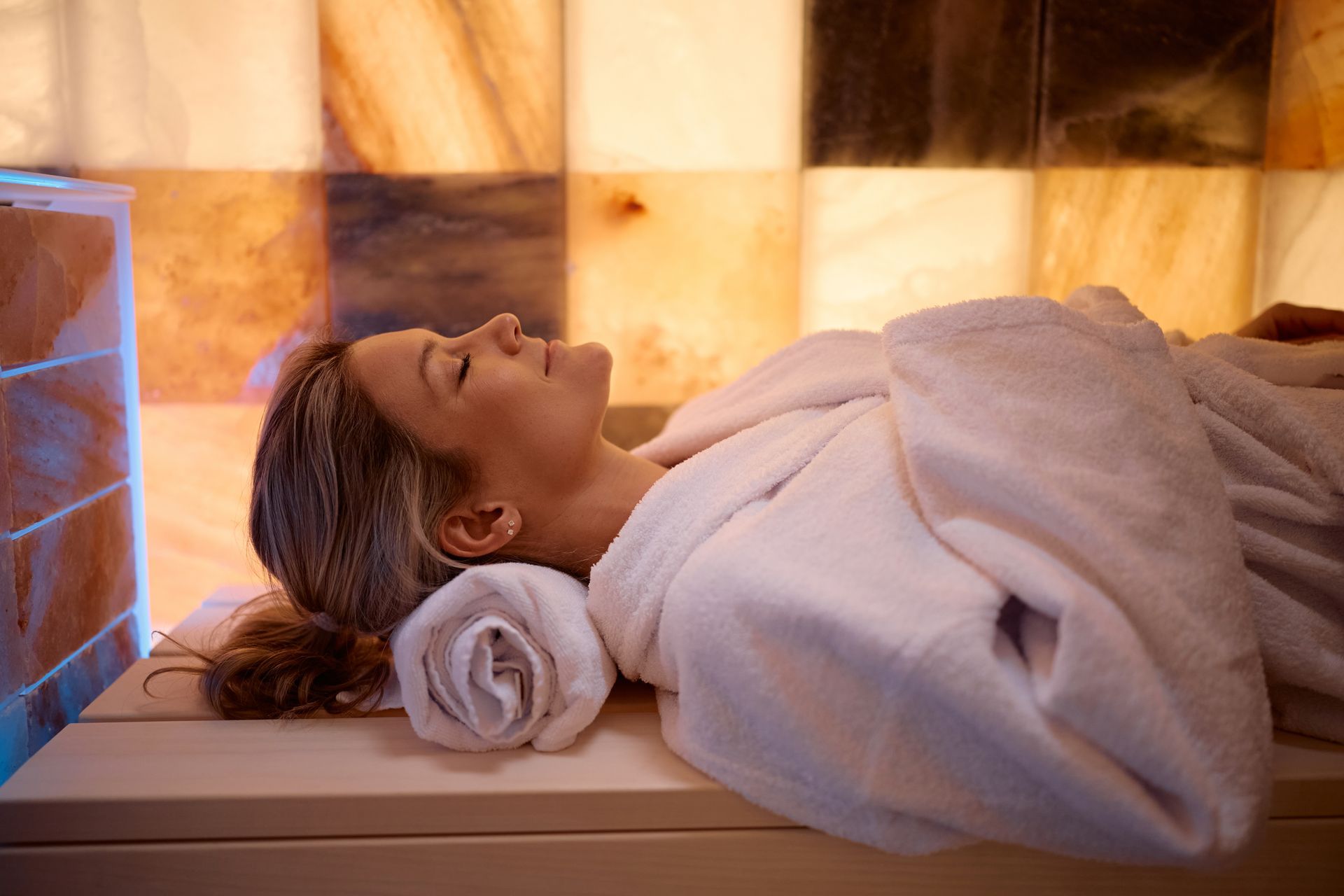 Person relaxing on a spa massage table with a rolled towel, warm amber-lit tiled room